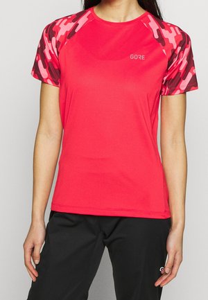Person trägt ein rotes Sportshirt mit kurzen Ärmeln mit rosa und dunkelroten Camouflage-Ärmeln und schwarze Hosen.