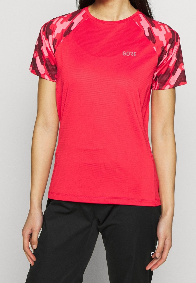 Personne portant un maillot de sport rouge à manches courtes avec des manches camouflage rose et rouge foncé et un pantalon noir.