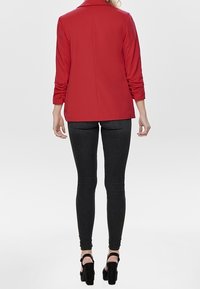 Blazer rojo con un corte ajustado, mangas largas y sutiles hombreras, combinado con jeans skinny negros y tacones negros con tiras.