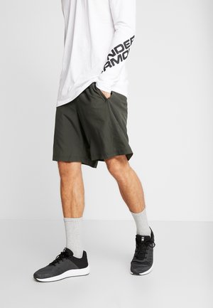 Homme portant un short de sport noir, un t-shirt blanc à manches longues avec un texte sur la manche, des chaussettes grises et des baskets noires, debout les mains dans les poches.