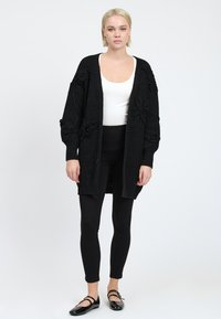 Zwarte gebreide cardigan met textuur van bloemdecoraties en open voorkant, gedragen over een witte top en gecombineerd met zwarte legging en flats.