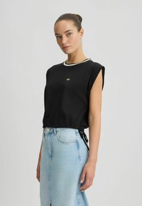Zwarte cropped top met een ronde halslijn, voorzien van een witte accentrand en een goudkleurig logo. Gecombineerd met een lichtblauwe denimrok.