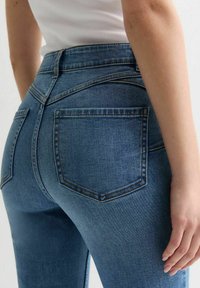 Jeans de mezclilla azul con cintura alta, dos bolsillos traseros y detalles de costura sutil, mostrados desde un ángulo lateral. Textura suave y diseño ajustado.