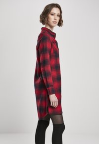 Robe chemise à carreaux rouges et noirs avec manches longues, devant à boutons, fentes sur les côtés et coupe confortable. Présente un col et une poche poitrine.