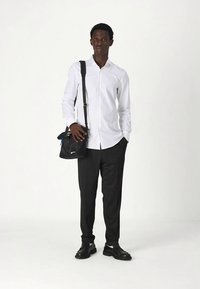 Biela košeľa s dlhým rukávom, čierne textúrované nohavice, čierna kabelka cez rameno a čierne topánky s bielymi akcentmi. Postoj v stoji na jednoduchom pozadí.