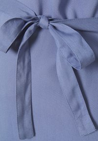 Tissu bleu clair avec une texture lisse mettant en valeur un détail de nœud noué à la taille, présentant de longs rubans fluides et des coutures soignées.