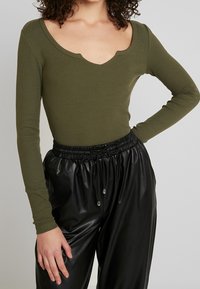 Olivgrüner, gerippter Langarm-Body mit tiefem V-Ausschnitt, kombiniert mit schwarzen Jogginghosen aus Kunstleder mit elastischem Bund.