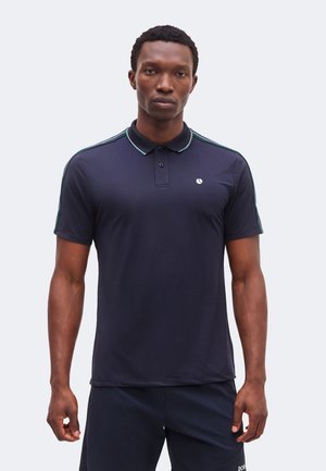 Άνδρας που φοράει σκούρο μπλε μπλουζάκι πόλο με κοντά μανίκια και λεπτές πράσινες και λευκές ρίγες στο γιακά και στα μανίκια, στέκεται μπροστά σε απλό φωτεινό φόντο.