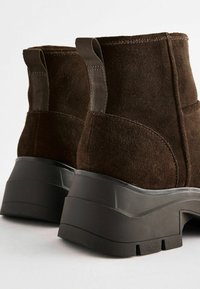 Bottes chelsea en suede marron avec une épaisse semelle en caoutchouc noir, dotées d'anses en boucle pour un enfilage facile et de détails de surpiqûres soignées.