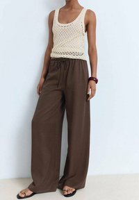 Jasnobeżowy szydełkowany top na ramiączkach w połączeniu z luźnymi brązowymi spodniami. Spodnie mają elastyczny pas i szeroki krój nogawek. Założone są płaskie sandały.