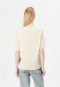 Majica svijetlo krem boje od pamuka s kratkim rukavima, okruglim izrezom i opuštenim krojem. Uparena s trapericama svijetloplave boje.