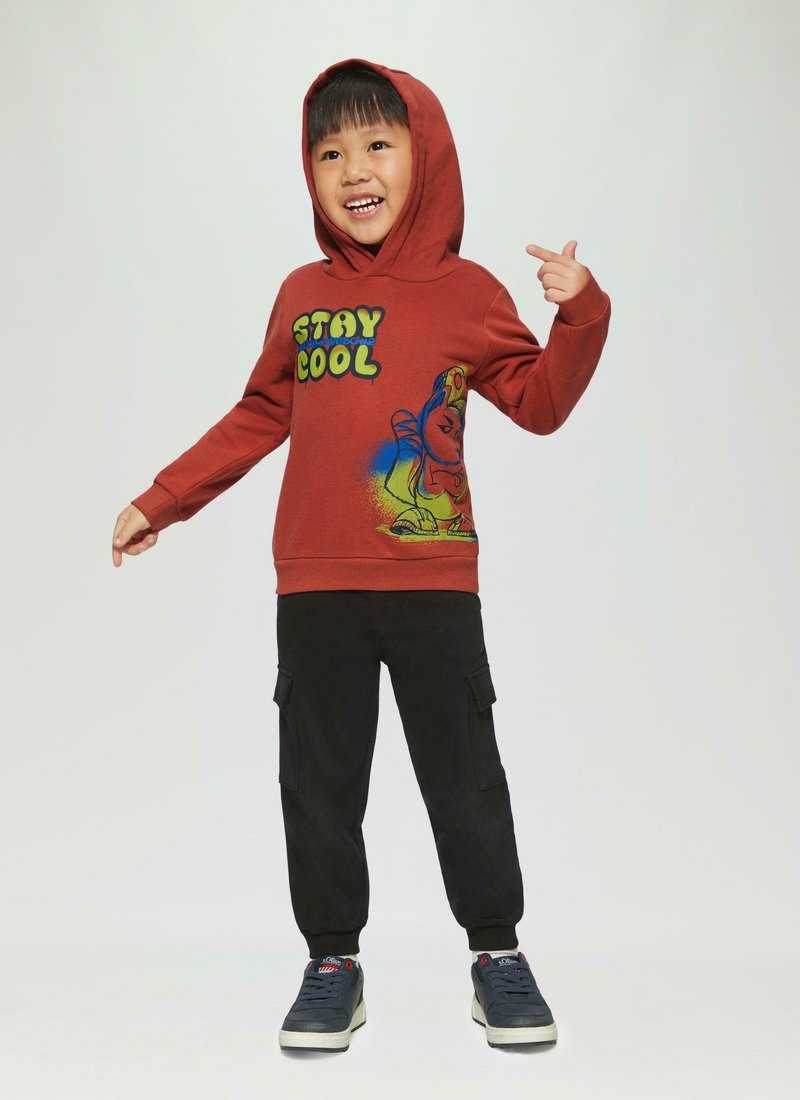 Hoodie rouge avec texte graphique "RESTEZ FROID" et design de dessin animé, associé à un pantalon cargo noir et des baskets bleu marine. Tissu lisse, coupe décontractée.