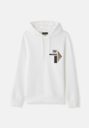 Haut à capuche blanc avec une poche kangourou, un cordon de serrage, et des patches graphiques en gris et noir sur la poitrine, fabriqué en tissu doux.