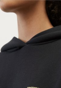 Černá mikina s výraznou přední kapsou, nastavitelnou šňůrkou a zlatým textovým detailem na oblasti hrudi. Hladká textura, casual design.