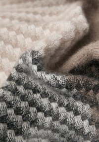 Gebreide plaid in een zachte mix van beige, grijs en zwart met een geruit patroon. Heeft een pluizige textuur en variërende dikte van de steken.