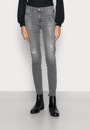 Osoba ubrana w szare, dopasowane, podniszczone dżinsy oraz czarne botki, stojąca na jasnym, jednolitym tle.