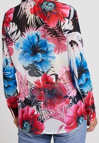 Blouse florale avec un design transparent présentant de grandes fleurs bleues, rouges et roses sur un fond noir, avec des détails en dentelle à la taille.
