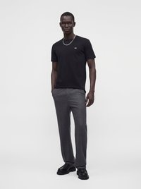 Homme debout portant un T-shirt noir, un pantalon gris, des chaussures noires et un collier chaîne argenté sur un fond blanc uni.