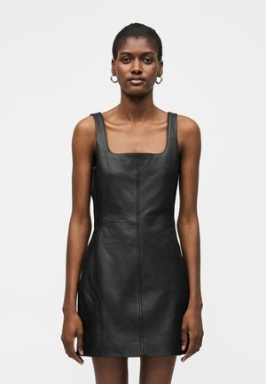 Kobieta o krótkich włosach, ubrana w czarną skórzaną mini sukienkę na ramiączkach z kwadratowym dekoltem i srebrne kolczyki w kształcie kół, stojąca na jednolitym tle.