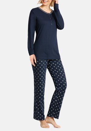 Frau trägt ein dunkelblaues Langarmshirt und dunkelblaue Pyjamahosen mit kleinem weißem Blumenmuster, steht barfuß vor einem schlichten Hintergrund.