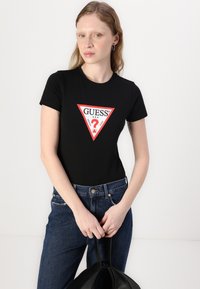 Camiseta de algodón negra con un gráfico de logo triangular rojo que incluye el texto "GUESS". Combinada con jeans de mezclilla azul oscuro y un bolso negro.