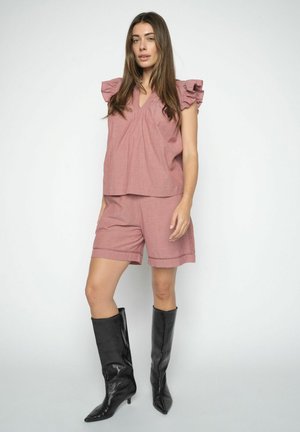 Kvinde iført en støvet lyserød top med flæser på ærmerne og matchende shorts samt sorte støvler med hæl, der går til midt på læggen, stående mod en ensfarvet baggrund.