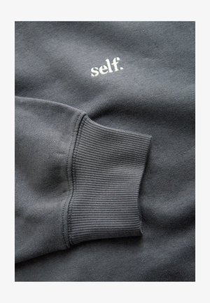 Mørkegrå sweatshirtærme med ribbet manchet og hvidt broderet tekst "self." på stoffet.