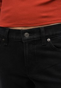 Gros plan sur la taille portant un jean en denim noir avec des boutons en métal et un haut côtelé orange brûlé rentré.