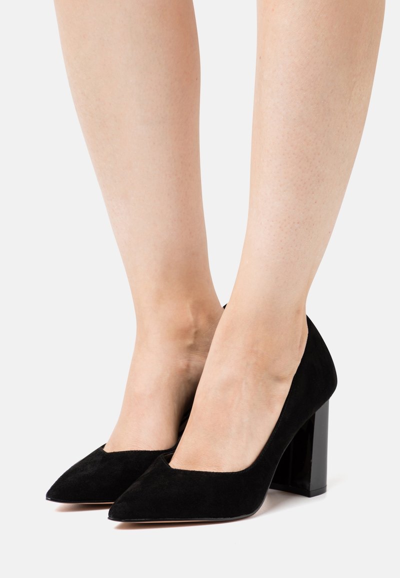 Escarpins en suède noir à talons hauts avec un bout pointu et un large talon carré brillant, présentant une texture lisse et un design minimaliste.