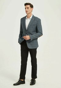 Blazer pour homme en tissu à carreaux bleus, avec revers crantés et bouton unique. Associé à une chemise blanche et un pantalon noir, terminé par des chaussures noires.