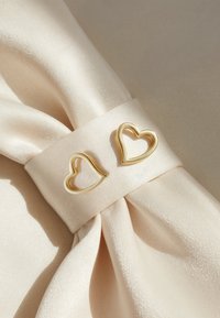 Guld hjärtformade klämmor på ett ljusbeige satin tyg med en jämn struktur och mjuk lyster, fästa vid basen av en båge.