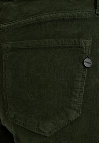 Zelený kordový textil s viditeľnými ryhami, obsahujúci náprsné vrecko a malú čiernu kovovú nálepku s označením "PIESZAK." Detail štruktúrovaného povrchu.