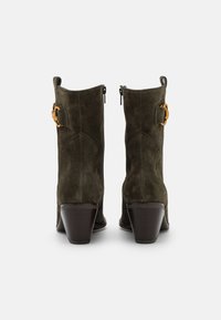 Stivaletti alla caviglia in suede verde oliva con un design curvato, dotati di una cerniera laterale, hardware dorato e un tacco in legno di altezza moderata.