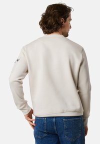 Felpa beige con scollatura rotonda, maniche lunghe e una tasca a toppa visibile sul braccio sinistro. Abbinata a jeans in denim blu.