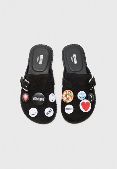 Deslizadores de ante negro con múltiples parches circulares que presentan gráficos variados, incluidos emojis y un diseño de pizza. Detalle de hebilla ajustable.