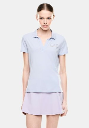 Giovane donna con capelli slicked back che indossa una polo celeste e una gonna lilla, in piedi davanti a uno sfondo bianco semplice.