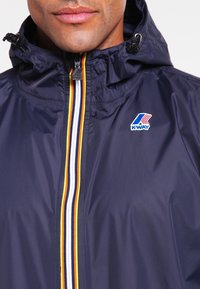 Laivastonsininen kevyt takki hupulla, jossa on etuvetoketju monivärisillä raidoilla ja pieni logo rinnassa.