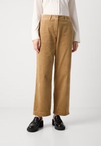 Beige wijdvallende corduroybroek gedragen met zwarte chunky-heeled loafers en een wit overhemd met lange mouwen, getoond van taille tot voeten.