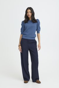 Femme aux cheveux foncés portant un pull à manches bouffantes bleu texturé, un pantalon large marine et des mocassins marron, debout sur fond blanc.