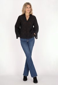 Schwarze Langarmbluse mit V-Ausschnitt und diamantartigem Ausschnittmuster, kombiniert mit blauen Schlagjeans und schwarzen Stiefeletten.