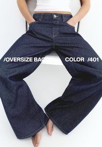 Sötét, túlméretezett bő farmernadrágot és fehér crop topot viselő személy, aki zsebeibe dugott kézzel, széttárt lábakkal ül.