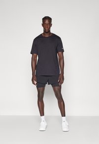 Schwarzes Kurzarm-T-Shirt mit rundem Ausschnitt, kombiniert mit schwarzen Shorts. Getragen mit weißen Sneakers und Knöchelsocken.