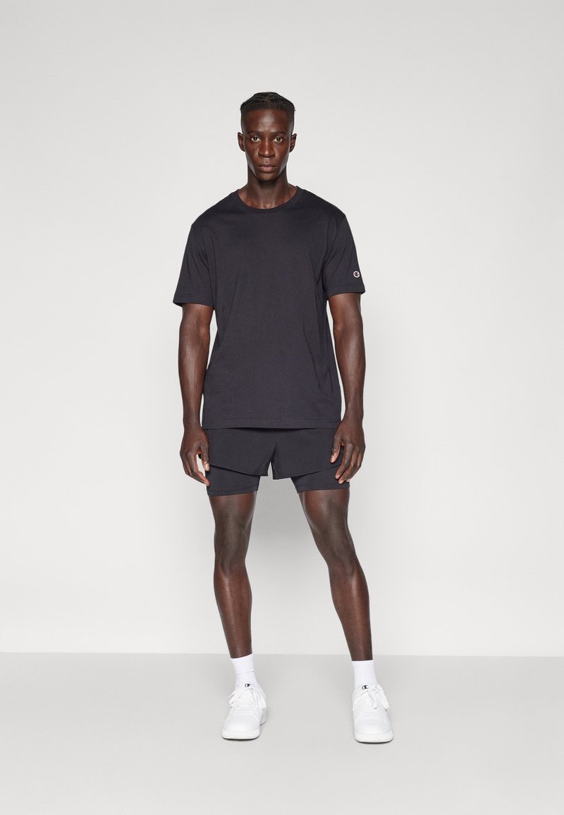Schwarzes Kurzarm-T-Shirt mit rundem Ausschnitt, kombiniert mit schwarzen Shorts. Getragen mit weißen Sneakers und Knöchelsocken.