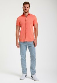Korte mouw koraalkleurige polo met een kraag, knoopsluiting en textiel textuur. Gecombineerd met lichtblauwe jeans en witte sneakers.