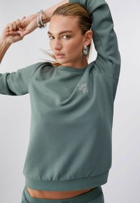 Sweat-shirt vert clair avec une coupe décontractée, poignets et ourlet côtelés. Présente un logo brodé subtil sur le devant. Texture lisse.