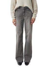 Kobieta ubrana w szare szerokie dżinsy, beżowy sweter z dzianiny oraz czarne, spiczaste buty stojąca na białym tle.