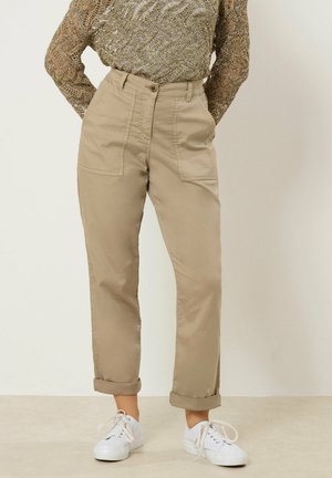 Person iført beige højttaljede bukser med opsmøgede kanter, sidelommer med lapper, hvide sneakers og en mønstret langærmet top, der er stoppet ind.