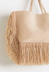 Sac tote beige en matériau tissé, avec un corps texturé orné de longues franges le long du bord inférieur et deux anses solides.