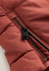 Bordeauxfarbene Pufferjacke aus Stoff mit schwarzem Reißverschluss, mit einer glatten Textur und gestepptem Design. Der Reißverschluss-Zug hat eine rechteckige Form.