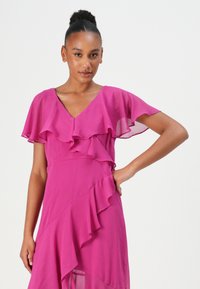 Robe fuchsia avec des manches flottantes et des volants en couches. Fabriquée en tissu léger, elle présente un décolleté en V et un ourlet asymétrique.
